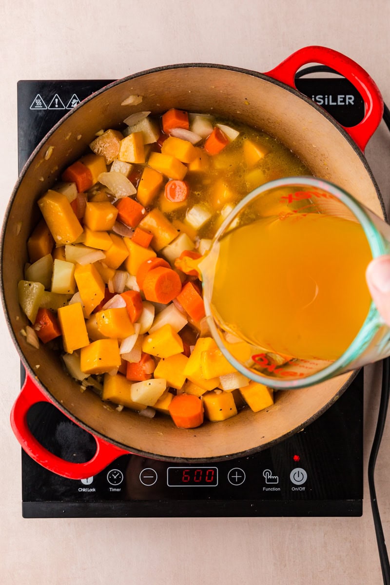 A pot on an induction stove holds chopped vegetables, like carrots and squash, simmering in liquid. A hand pours extra broth from a measuring cup into the pot, perfecting the butternut squash and carrot soup. The stove display indicates a temperature setting of 600.