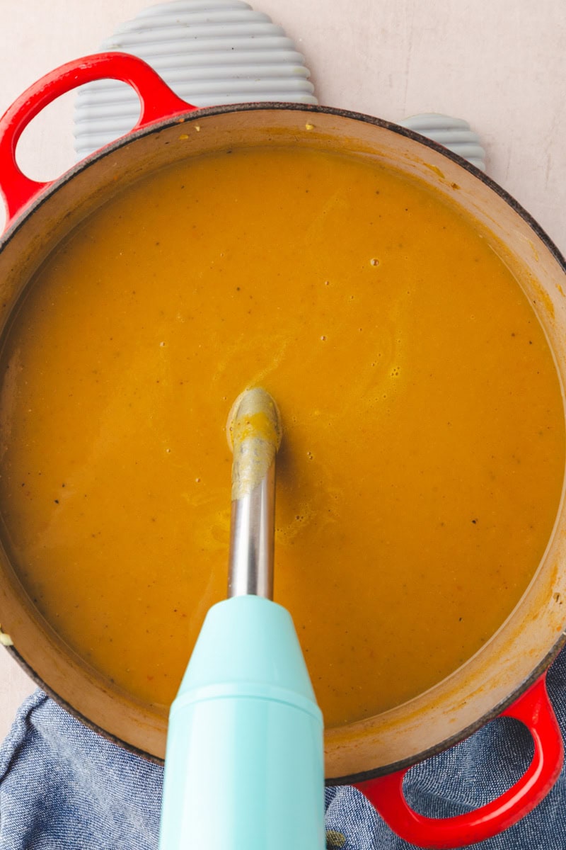 An immersion blender is blending a pot of creamy butternut squash and carrot soup in a red pot placed on a blue towel. The soup appears smooth and thick, suggesting it's a pureed vegetable delight.