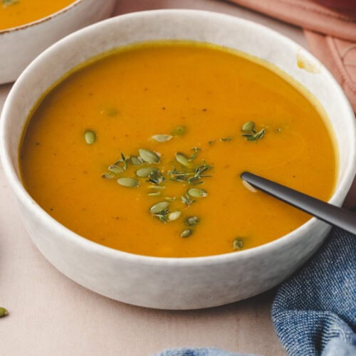 A bowl of creamy butternut squash and pumpkin soup garnished with pumpkin seeds and fresh herbs. A black spoon rests in the bowl, and a textured blue cloth is partially visible nearby.