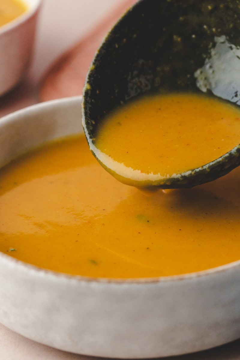 Close-up of a ladle pouring creamy butternut squash and carrot soup into a white bowl. The soup appears smooth and thick, with a rich, warm color. A blurred backdrop hints at more bowls and a soft surface.