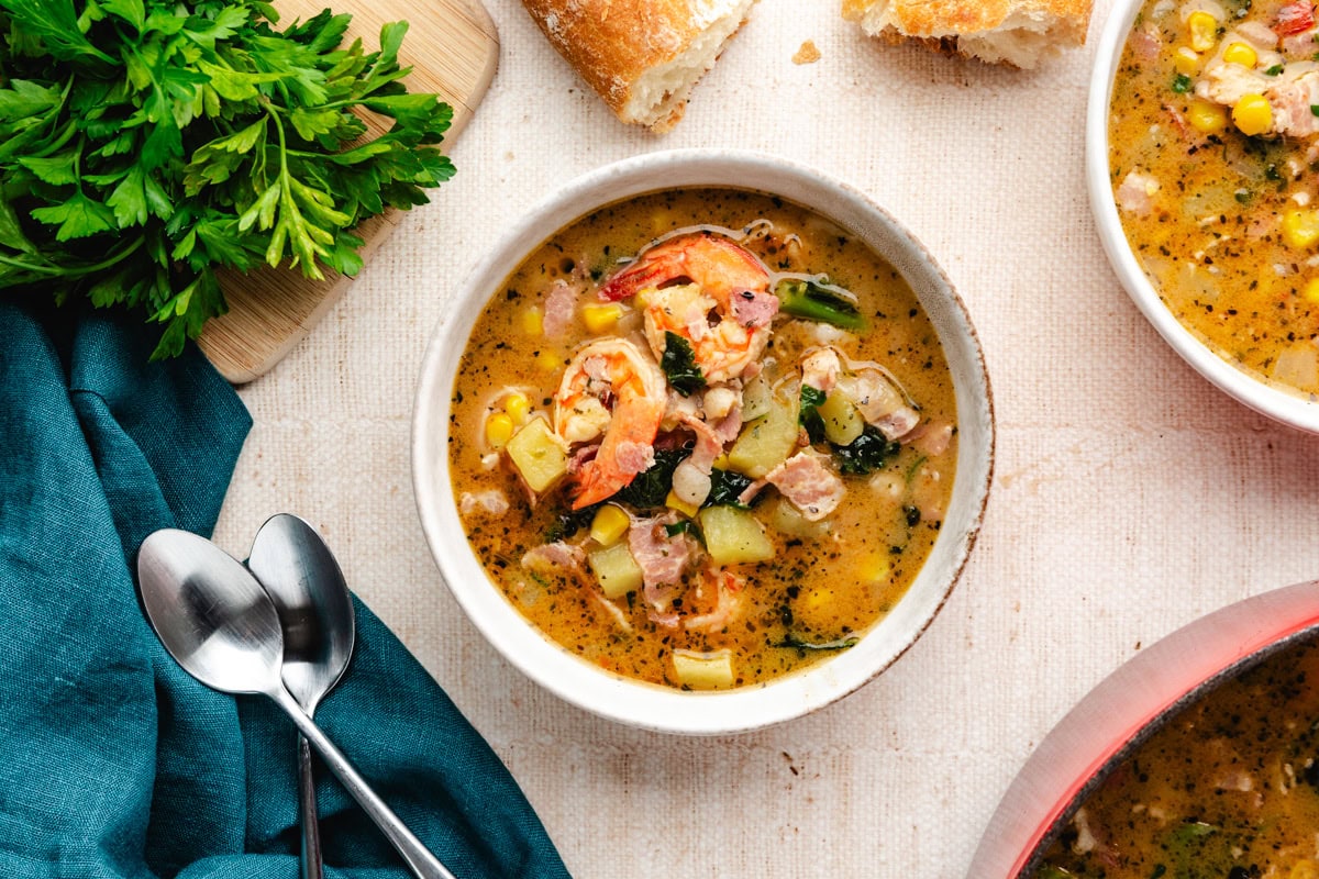 A bowl of hearty seafood chowder filled with shrimp, vegetables, and herbs. Next to the bowl are two spoons, fresh parsley on a cutting board, a blue napkin, and pieces of crusty bread. Another partial bowl of chowder is visible in the corner.