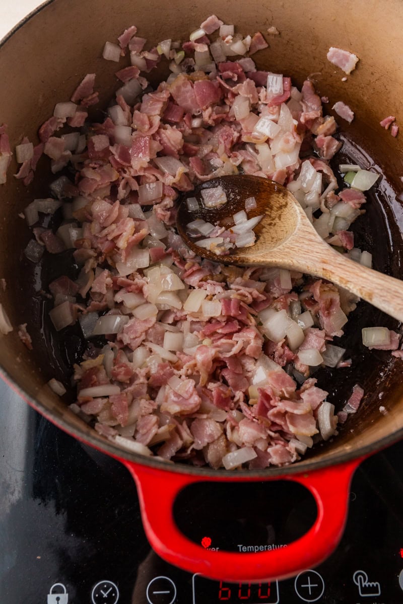 Chopped onions and bacon are cooking in a red pot on a stovetop, forming the savory base for a hearty corn and shrimp chowder. A wooden spoon rests in the pot, partially submerged in the sizzling ingredients.