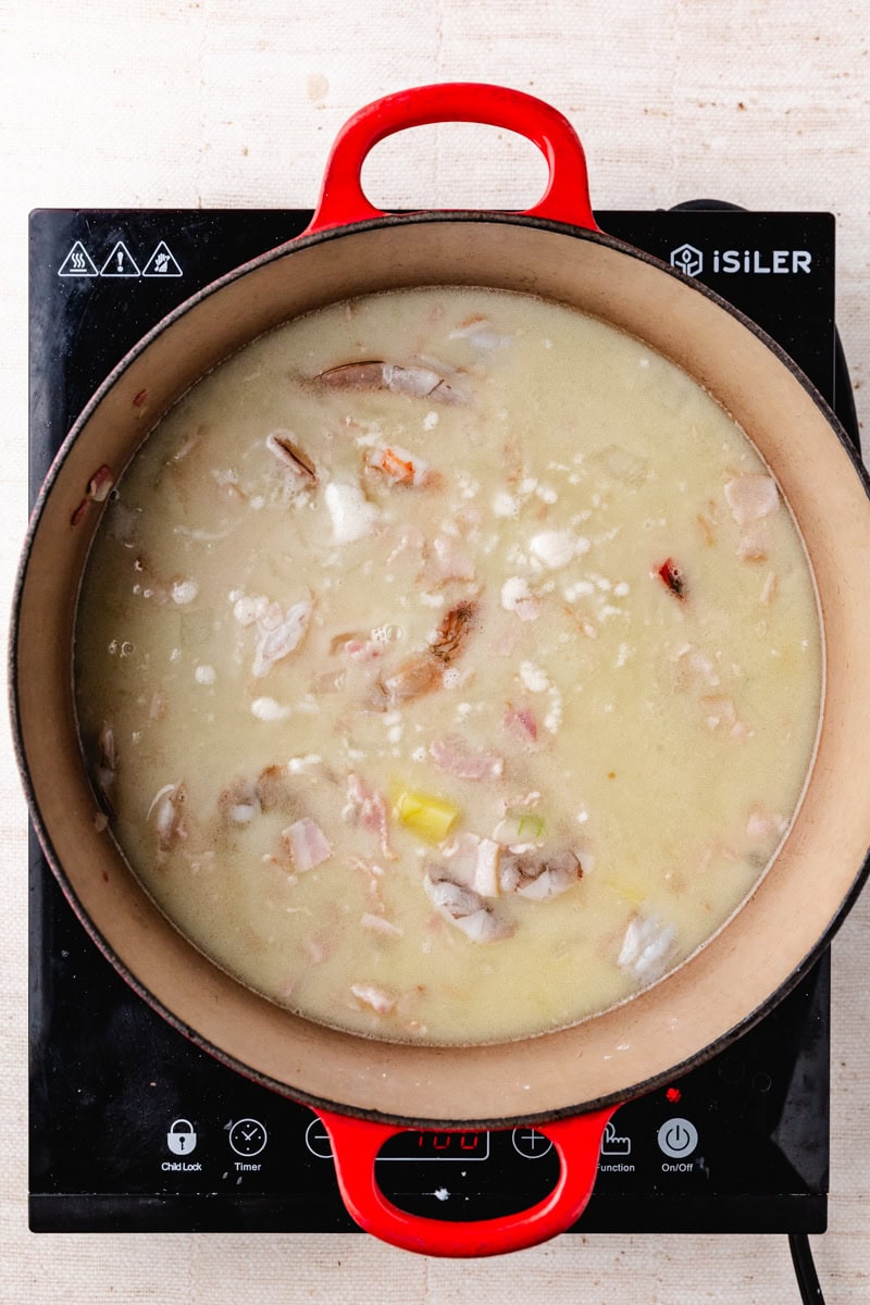 A red pot on a black induction cooktop contains a creamy corn and shrimp chowder with chunks of chicken, leeks, and pieces of bacon. The soup is simmering gently, and the stove surface shows a digital display.