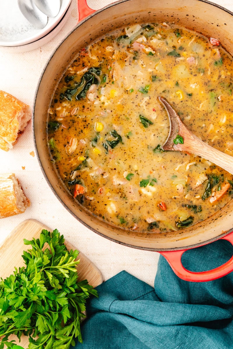 A hearty vegetable soup reminiscent of a corn chowder sits in a red pot with a wooden spoon. It brims with corn, beans, spinach, and tomatoes, subtly seasoned with herbs. Fresh parsley and bread are on the side, complementing the blue cloth and silver serving spoons nearby on the table.