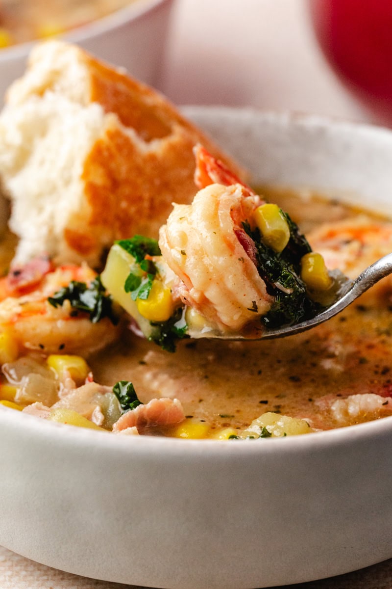 Close-up of a bowl of creamy corn and shrimp chowder filled with greens and chunks of bread. A spoon holds a piece of shrimp, showcasing the dish's savory ingredients and rich texture.
