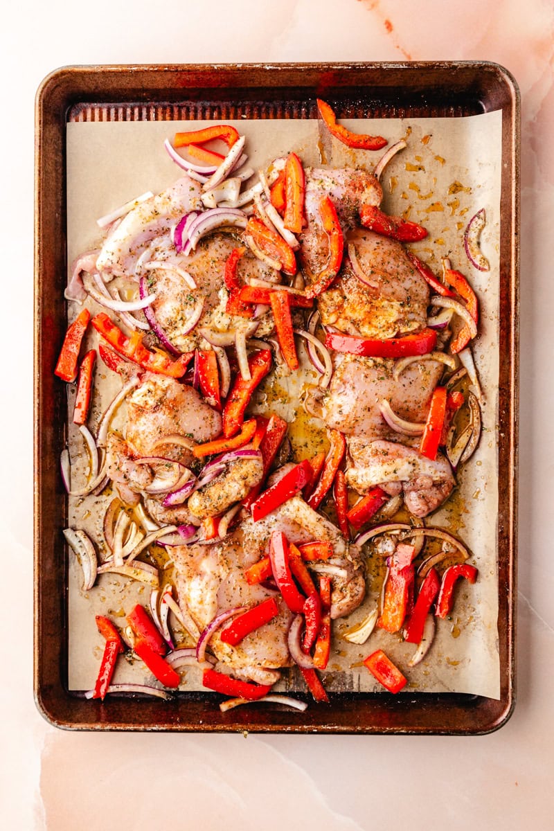 Italian baked chicken thighs rest on a baking sheet alongside sliced red bell peppers and onions, seasoned to perfection. These flavorful ingredients are artfully arranged on parchment paper, ready for roasting.