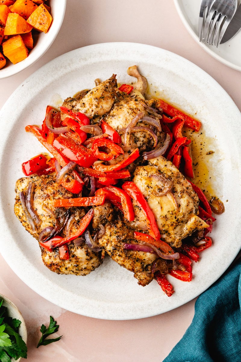 A white plate featuring Italian baked chicken thighs, topped with sautéed red bell peppers and onions. The dish is accompanied by a bowl of roasted sweet potatoes and a fork on a napkin. The setting is on a light pink surface, adding charm to the presentation.