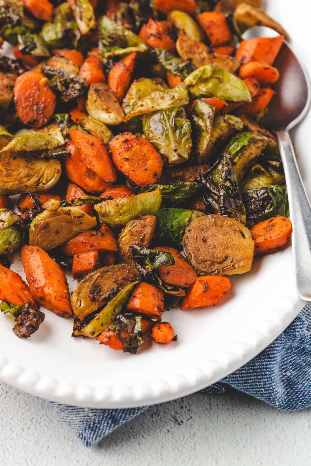 A close-up of a white plate filled with oven-roasted carrots and Brussels sprouts. The vegetables are deliciously seasoned, with a serving spoon resting on the plate. A blue cloth is partially visible under the plate.