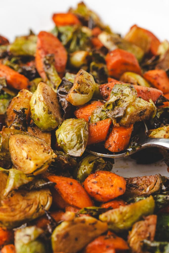 Close-up of roasted carrots and brussels sprouts. The vegetables are seasoned with herbs and spices, creating a colorful and appetizing dish. A spoon is partially visible, suggesting serving.
