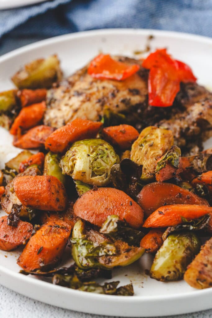 A plate of roasted carrots and Brussels sprouts accompanies a piece of seasoned meat, possibly chicken. The vibrant dish is beautifully arranged on a white plate with a blue cloth in the background, showcasing a harmonious blend of flavors and colors.