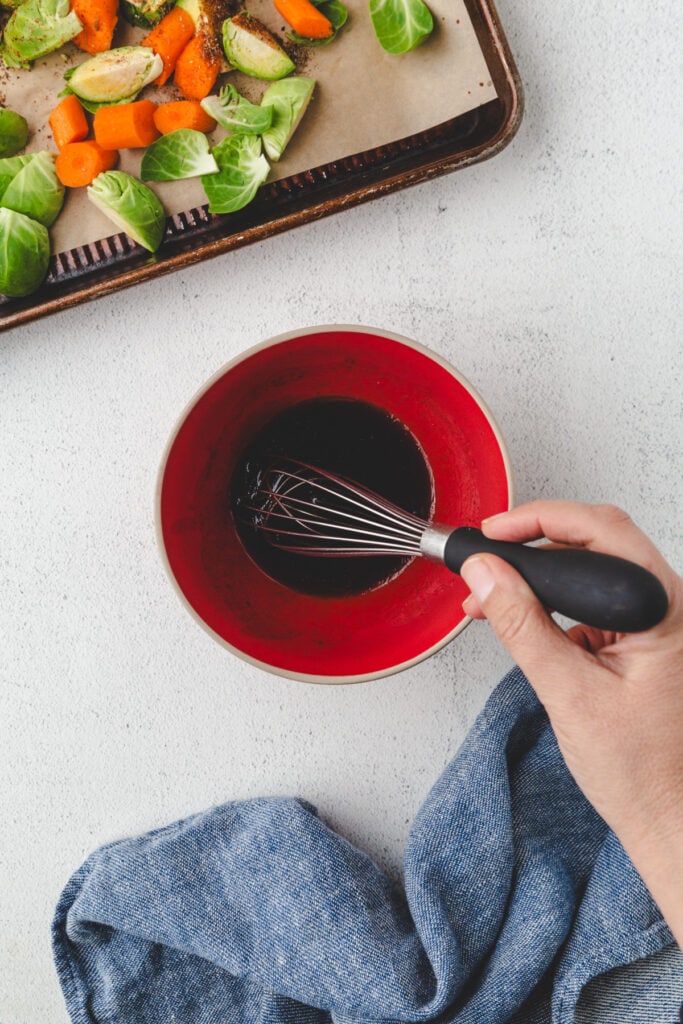 A person whisks a dark liquid in a red bowl, preparing to coat the baking tray filled with roasted carrots and Brussels sprouts. A blue kitchen towel rests on the white countertop, adding a pop of color to the culinary scene.