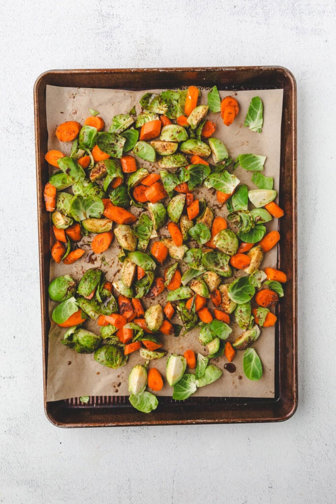 A baking tray with roasted carrots and Brussels sprouts, perfectly seasoned with herbs, rests on parchment paper. The vibrant vegetables are cut into pieces and evenly spread across the tray against a light background.