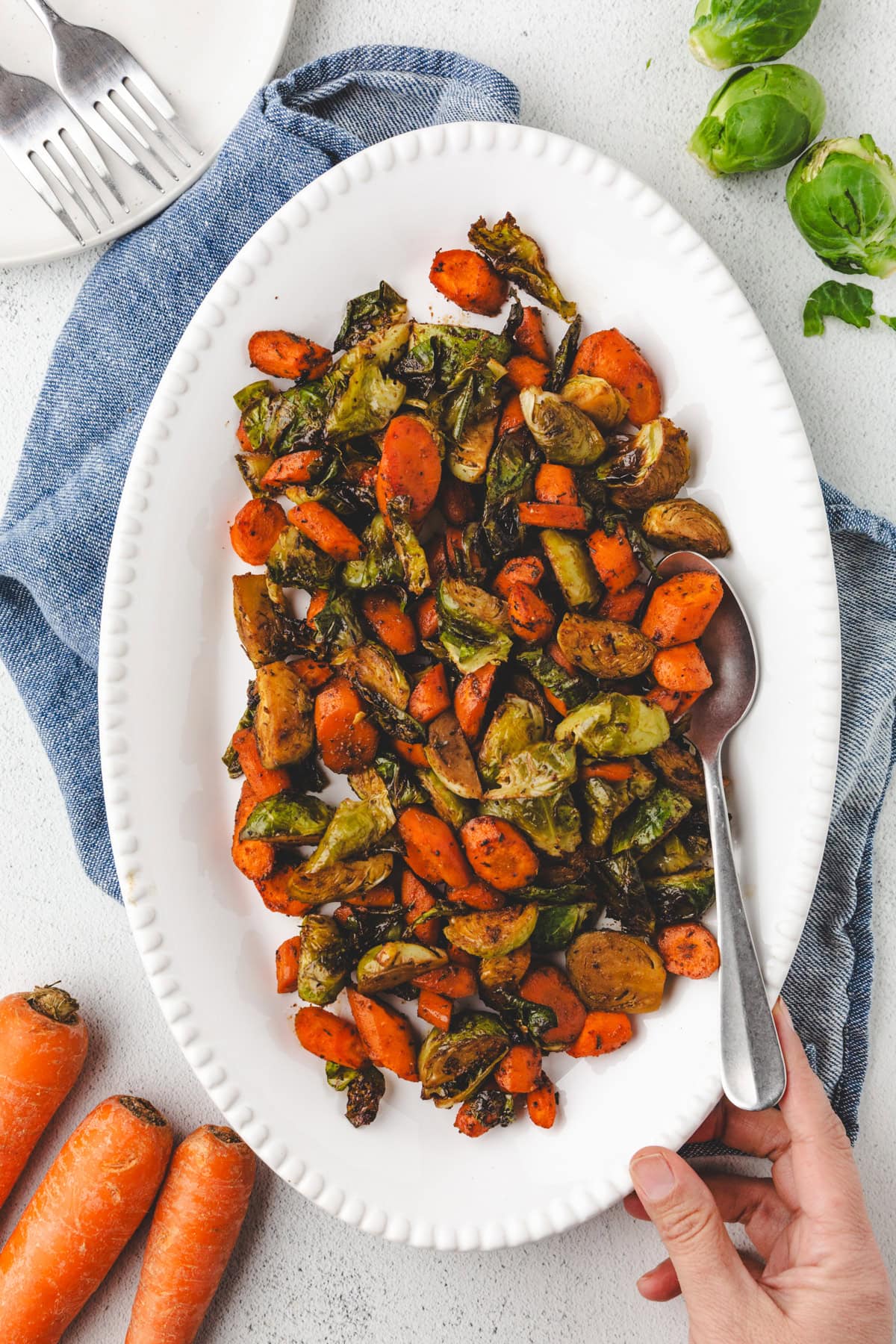 A hand holds a white oval plate with roasted Brussels sprouts, carrots, and other vegetables. The plate rests on a blue cloth, surrounded by whole carrots, a fork, and a few Brussels sprouts on a light surface.