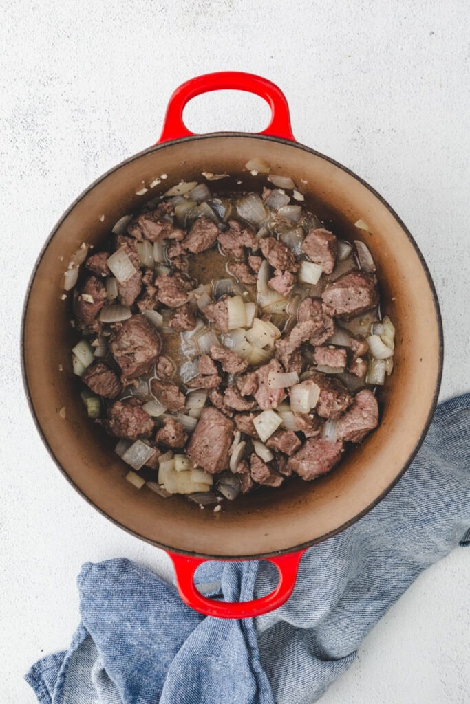 A red pot filled with savory Whole30 beef stew, simmering with pieces of beef and chopped onions, rests on a light surface beside a crumpled blue cloth.