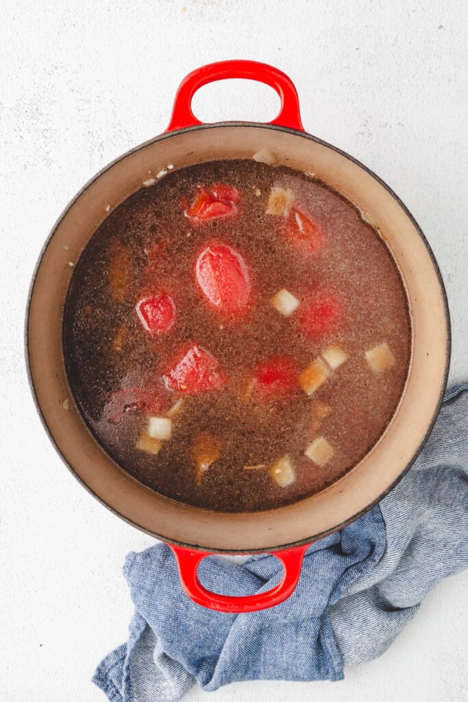A red pot filled with a hearty Whole30 beef stew, brimming with chunks of tomatoes, onions, and other vegetables. The pot rests on a white surface, partially wrapped in a blue cloth for a cozy touch.
