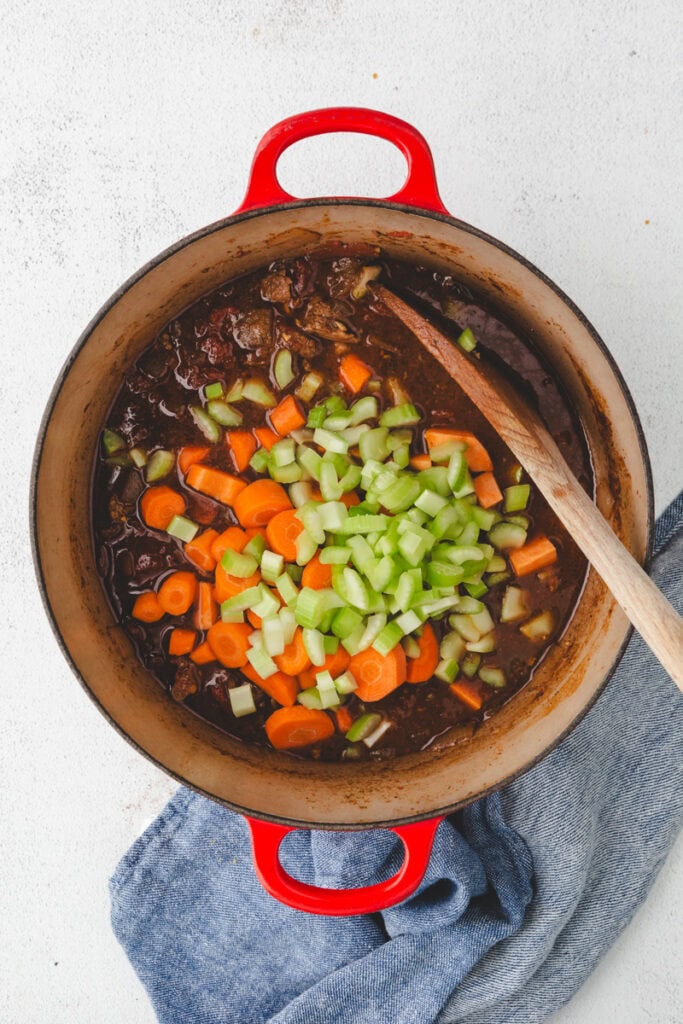 A red pot filled with a hearty Whole30 beef stew, topped with chopped carrots and celery, sits on a light surface. A wooden spoon rests inside the pot, and a blue cloth is nearby.