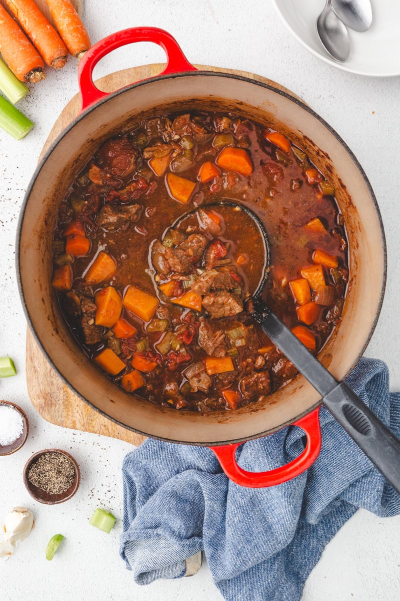 A red pot brims with Whole30 beef stew, showcasing tender chunks of beef, carrots, and celery in a rich broth. A ladle rests nearby. Salt, pepper, garlic cloves, carrots, and celery are scattered on a white surface accentuated by a blue cloth.