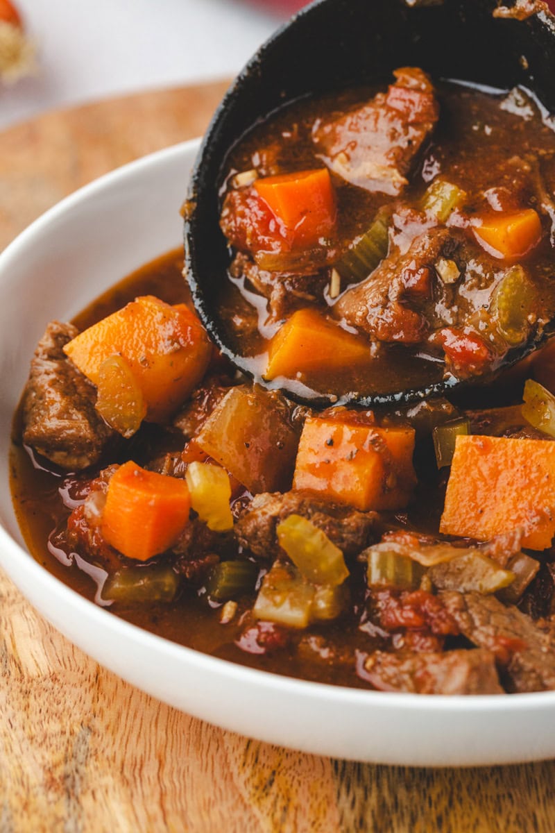 A close-up of Whole30 beef stew in a white bowl, featuring chunks of tender beef, carrots, and celery in a rich, dark broth. A ladle serves the savory stew, emphasizing its thick and hearty texture.