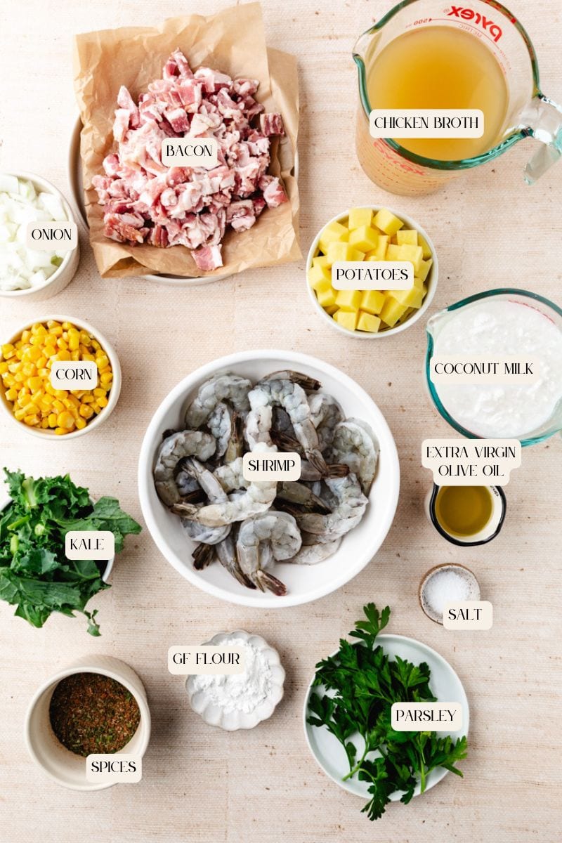 Ingredients for corn and shrimp chowder laid out on a table. Includes shrimp, bacon, kale, onion, corn, potatoes, chicken broth, coconut milk, GF flour, spices, extra virgin olive oil, salt, and parsley. Labels indicate each item clearly.