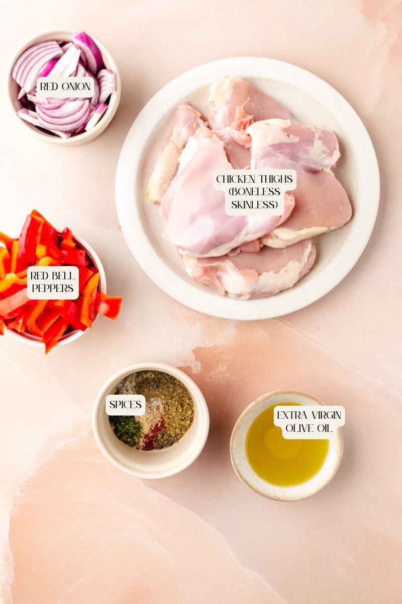 Ingredients on a marble surface: a plate of boneless, skinless chicken thighs ready for Italian baked chicken thighs, bowls of red onion slices, red bell pepper slices, mixed spices, and a dish of extra virgin olive oil.