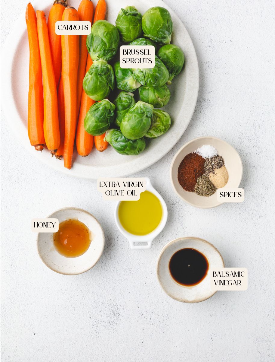 Roasted carrots and Brussels sprouts rest on a plate, encircled by bowls of extra virgin olive oil, honey, balsamic vinegar, and a mix of spices on a light surface. Ingredients are neatly labeled.