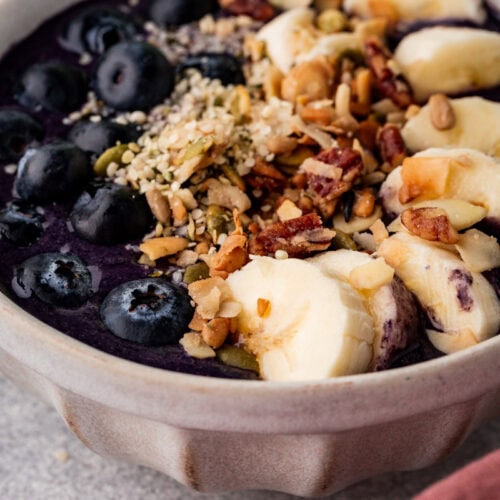 A ceramic bowl filled with a purple smoothie, topped with sliced bananas, blueberries, nuts, seeds, and granola. The bowl is placed on a textured surface with a hint of a pink cloth visible in the corner.