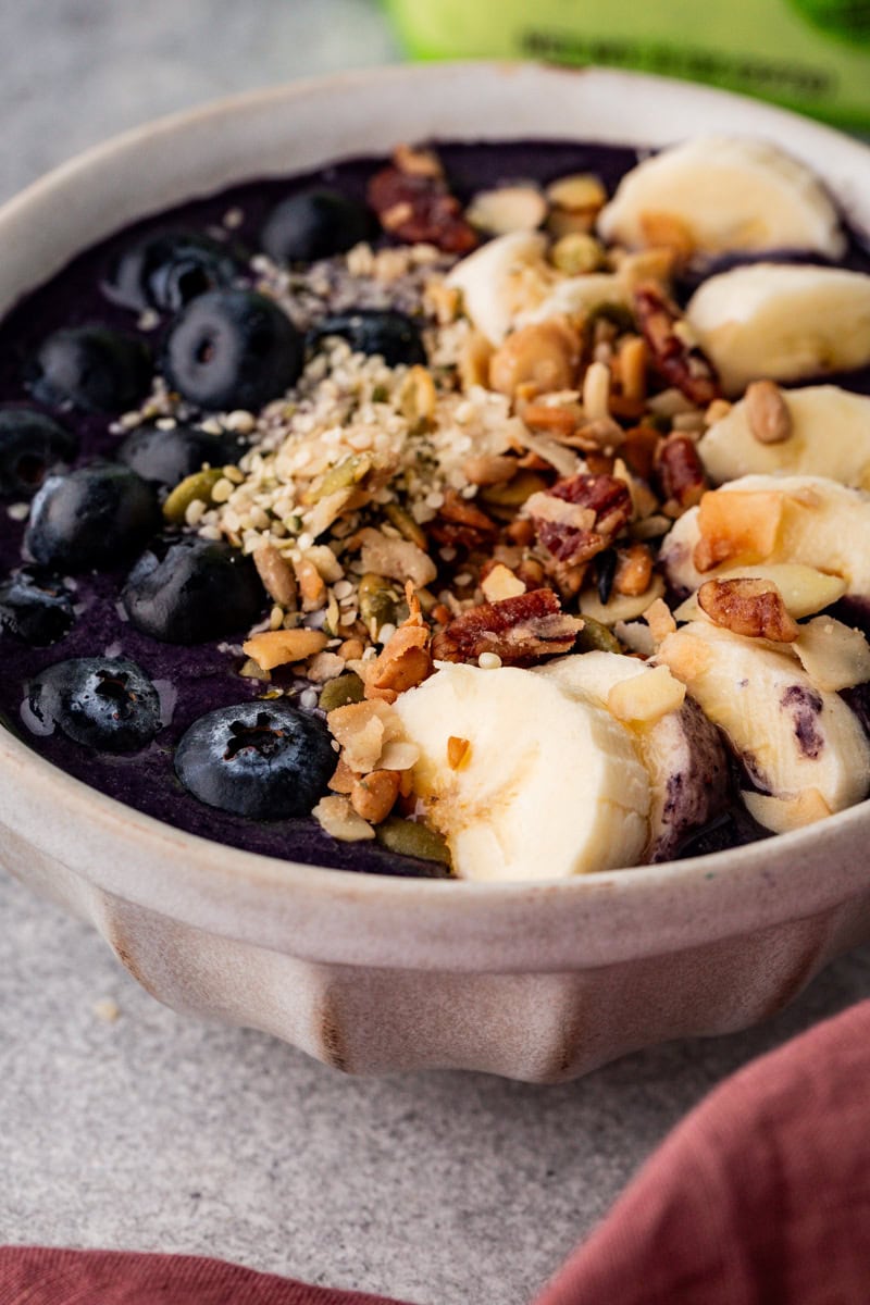 A ceramic bowl filled with a purple smoothie, topped with sliced bananas, blueberries, nuts, seeds, and granola. The bowl is placed on a textured surface with a hint of a pink cloth visible in the corner.