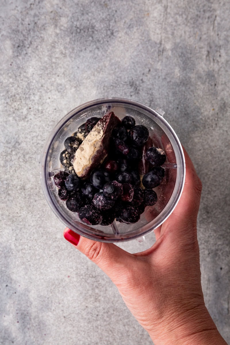 A hand with red nail polish holds a clear cup filled with frozen blueberries and a piece of frozen banana against a gray background.