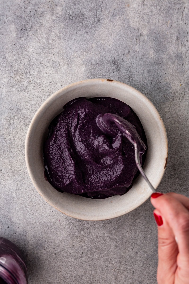 A hand with red nail polish holds a spoon, stirring a smooth, dark purple paste in a white bowl on a gray surface.