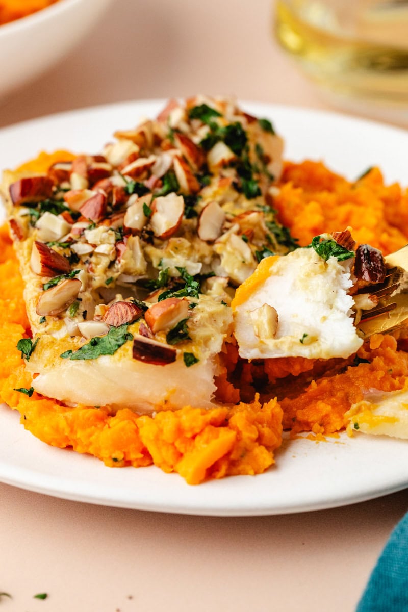 A dish of almond-crusted cod topped with herbs, served on a bed of mashed sweet potatoes. A forkful of the fish is lifted, revealing its flaky texture. A glass of white wine is visible in the background, complementing the meal perfectly.