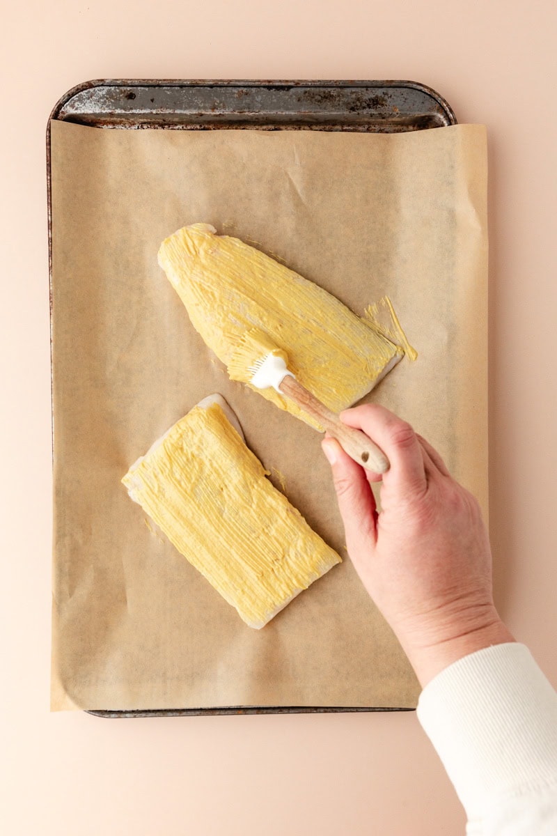 A person is brushing a mustard-like spread onto two almond-crusted pieces of cod on a parchment-lined baking sheet. The background is a neutral shade, and the hand is holding a wooden brush.