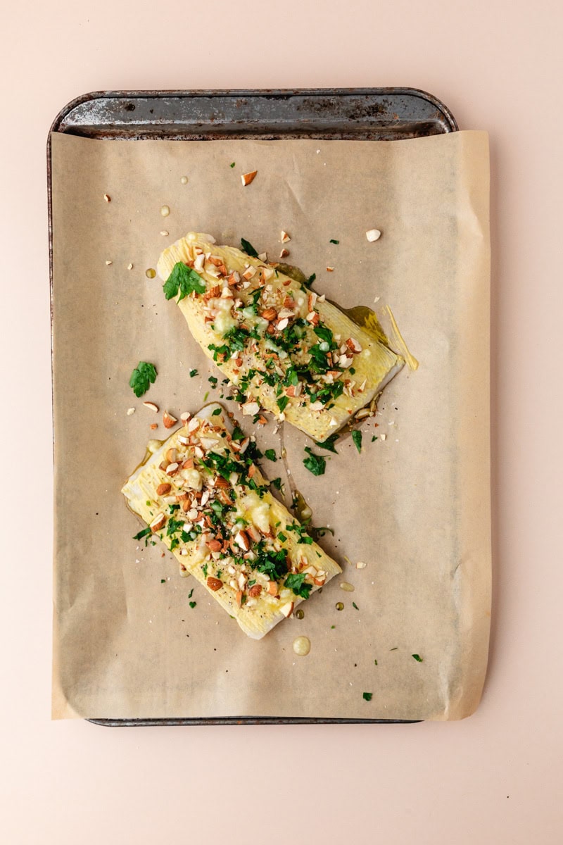 Two slices of bread topped with a delightful almond crusted cod, sprinkled with chopped almonds and fresh herbs, are drizzled with olive oil on a parchment-lined baking sheet.