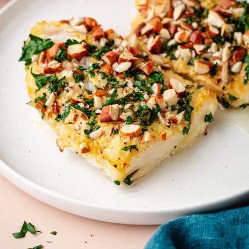 Two pieces of white fish topped with chopped almonds and herbs on a white plate. A blue cloth partially visible in the corner adds a pop of color. The dish is set against a soft, light background.