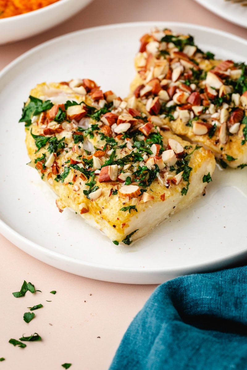 Two pieces of white fish topped with chopped almonds and herbs on a white plate. A blue cloth partially visible in the corner adds a pop of color. The dish is set against a soft, light background.