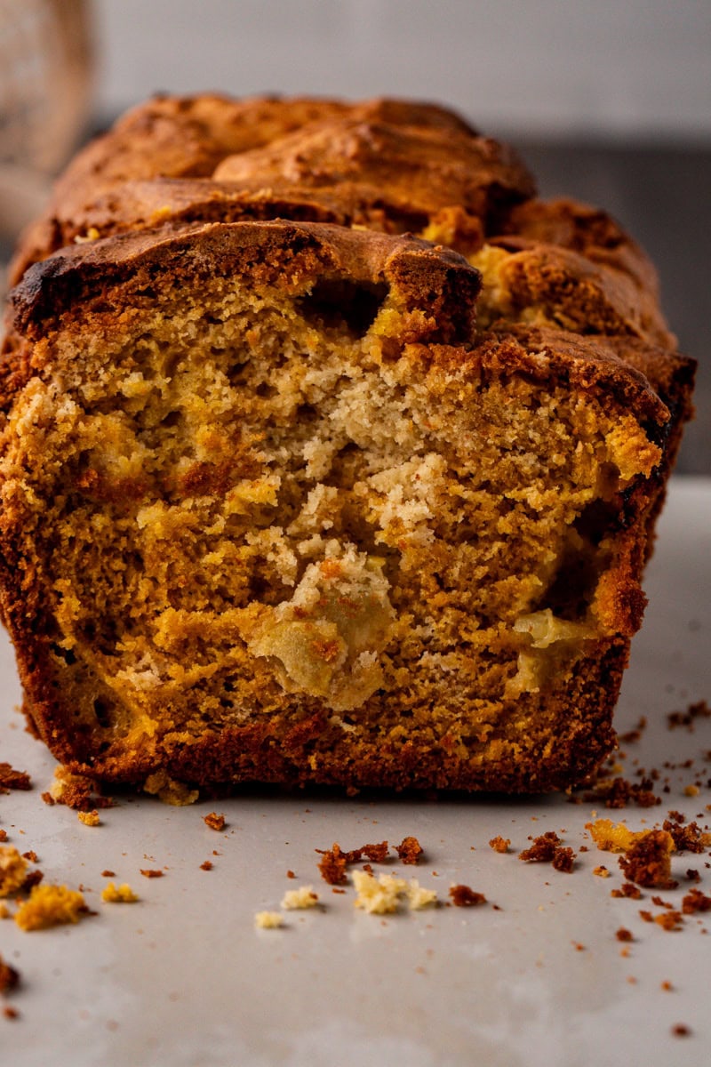 Close-up of a loaf of banana bread with a golden-brown crust, showcasing a moist and crumbly texture reminiscent of apple pumpkin bread. Scattered crumbs adorn the cutting surface, while visible banana pieces enhance its homemade charm.
