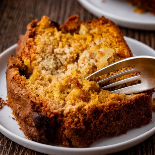 A close-up of a slice of crumbly apple cake on a white plate with a fork digging into it. The cake, with a golden-brown crust, sits on a rustic wooden table, highlighting its moist interior texture. Another slice is partially visible in the background, resembling spiced pumpkin bread.