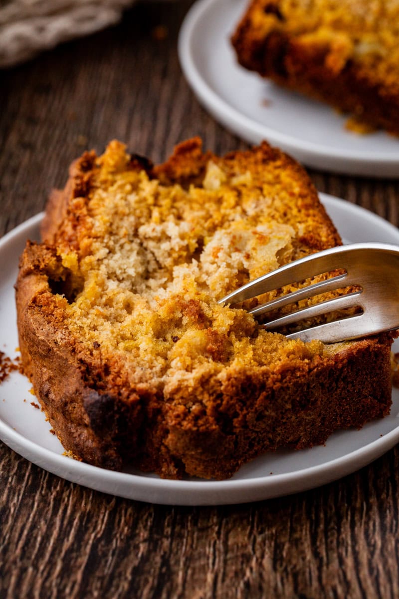 A close-up of a slice of crumbly apple cake on a white plate with a fork digging into it. The cake, with a golden-brown crust, sits on a rustic wooden table, highlighting its moist interior texture. Another slice is partially visible in the background, resembling spiced pumpkin bread.