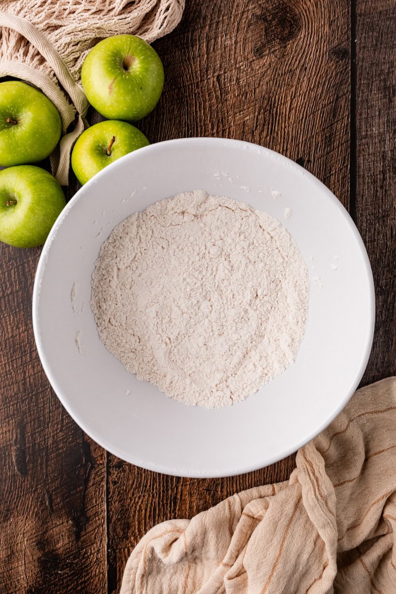 A white bowl with a flour mixture rests on a wooden surface, surrounded by a few green apples, hinting at the rustic charm of apple pumpkin bread. A beige cloth adds a touch of warmth to this cozy baking scene.