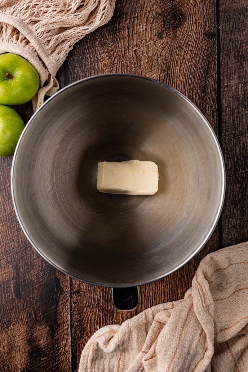 A metal mixing bowl with a stick of butter sits on a wooden surface, ready for baking apple pumpkin bread. Green apples surround the scene, alongside a beige cloth. A mesh bag peeks in from the top left corner, hinting at more autumn flavors to come.