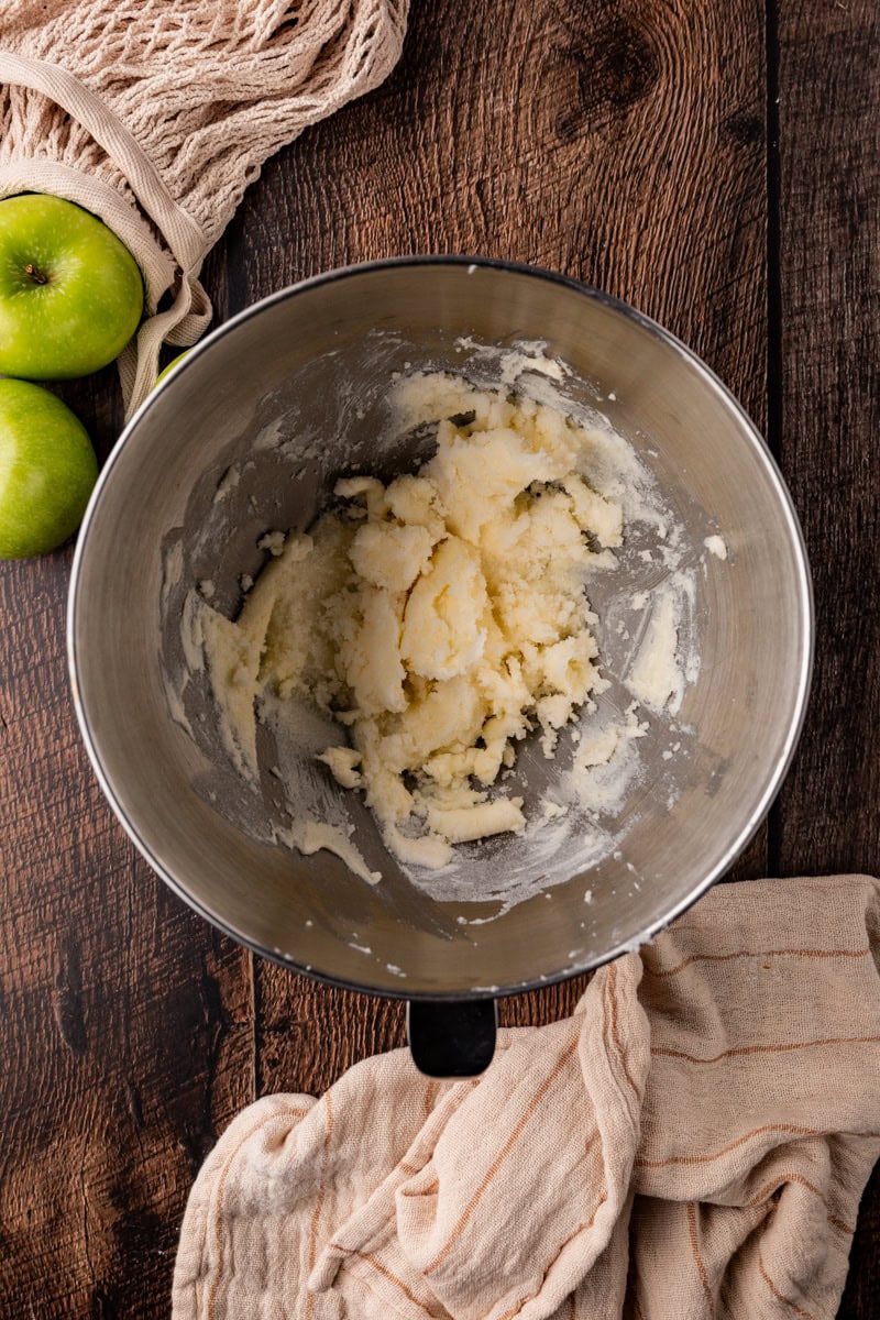 A stainless steel mixing bowl contains a creamy butter and sugar mixture. The bowl sits on a rustic wooden surface, accompanied by green apples, hinting at the aroma of freshly baked apple pumpkin bread. A beige kitchen cloth and a mesh produce bag are partially visible nearby.