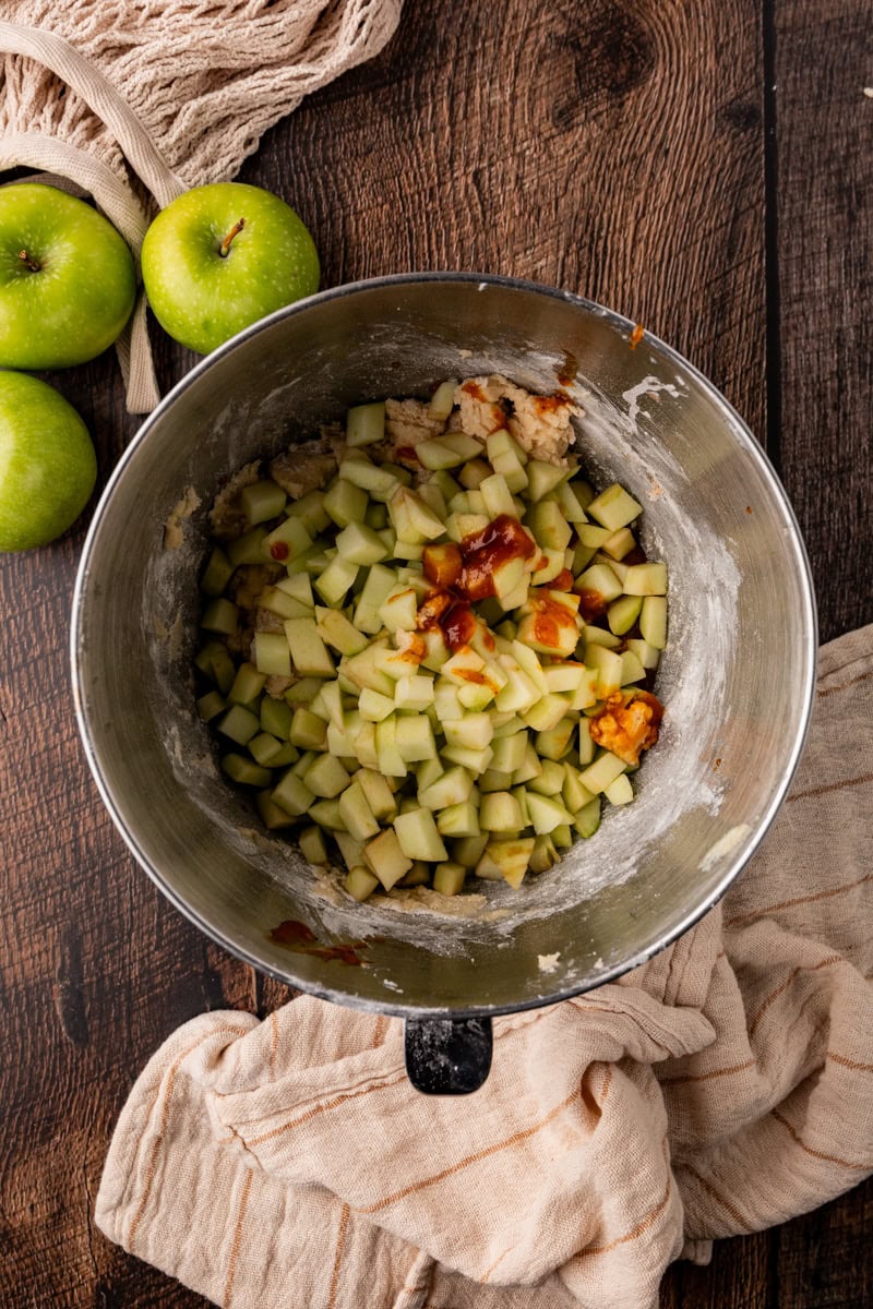 A metal mixing bowl filled with diced green apples, cinnamon, and other ingredients sits on a wooden surface, evoking the cozy aroma of apple pumpkin bread. Fresh green apples and a beige cloth are nearby.