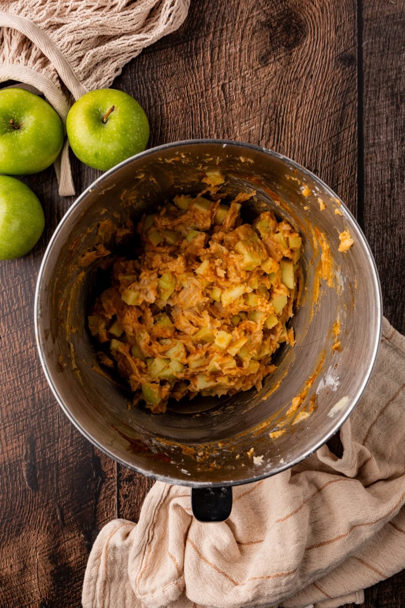 A mixing bowl with apple pie filling sits on a rustic wooden surface, evoking the cozy charm of apple pumpkin bread. Two fresh green apples and a beige cloth accompany the scene, enhancing its warm, inviting appeal.