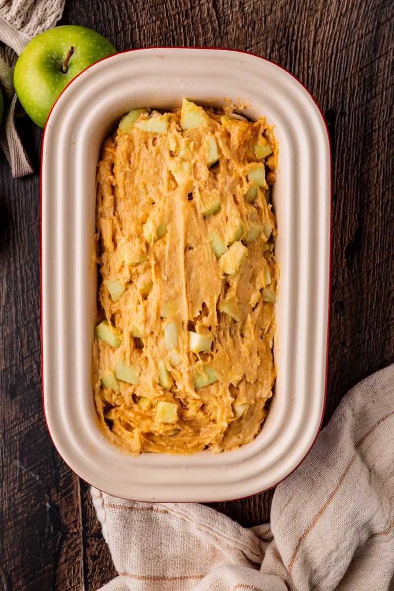 A baking dish filled with raw apple cake batter, reminiscent of apple pumpkin bread, topped with apple chunks on a wooden table. A green apple and a beige cloth are also visible beside the dish.