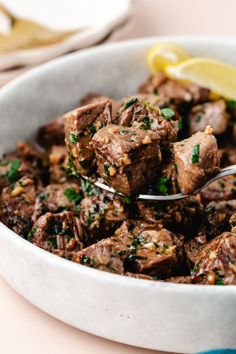 A bowl of Mediterranean garlic butter steak bites garnished with chopped parsley. A spoon holds a piece of the seasoned steak, highlighting its juicy texture. A lemon wedge is visible in the background for added flavor.