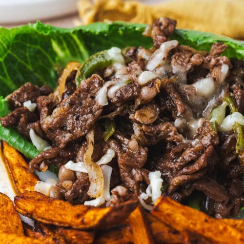 Close-up of a dish reminiscent of classic Philly cheesesteaks, featuring juicy beef strips with melted cheese and sautéed onions on a bed of lettuce, accompanied by seasoned, crispy sweet potato fries. The vibrant colors and textures create an appetizing presentation.