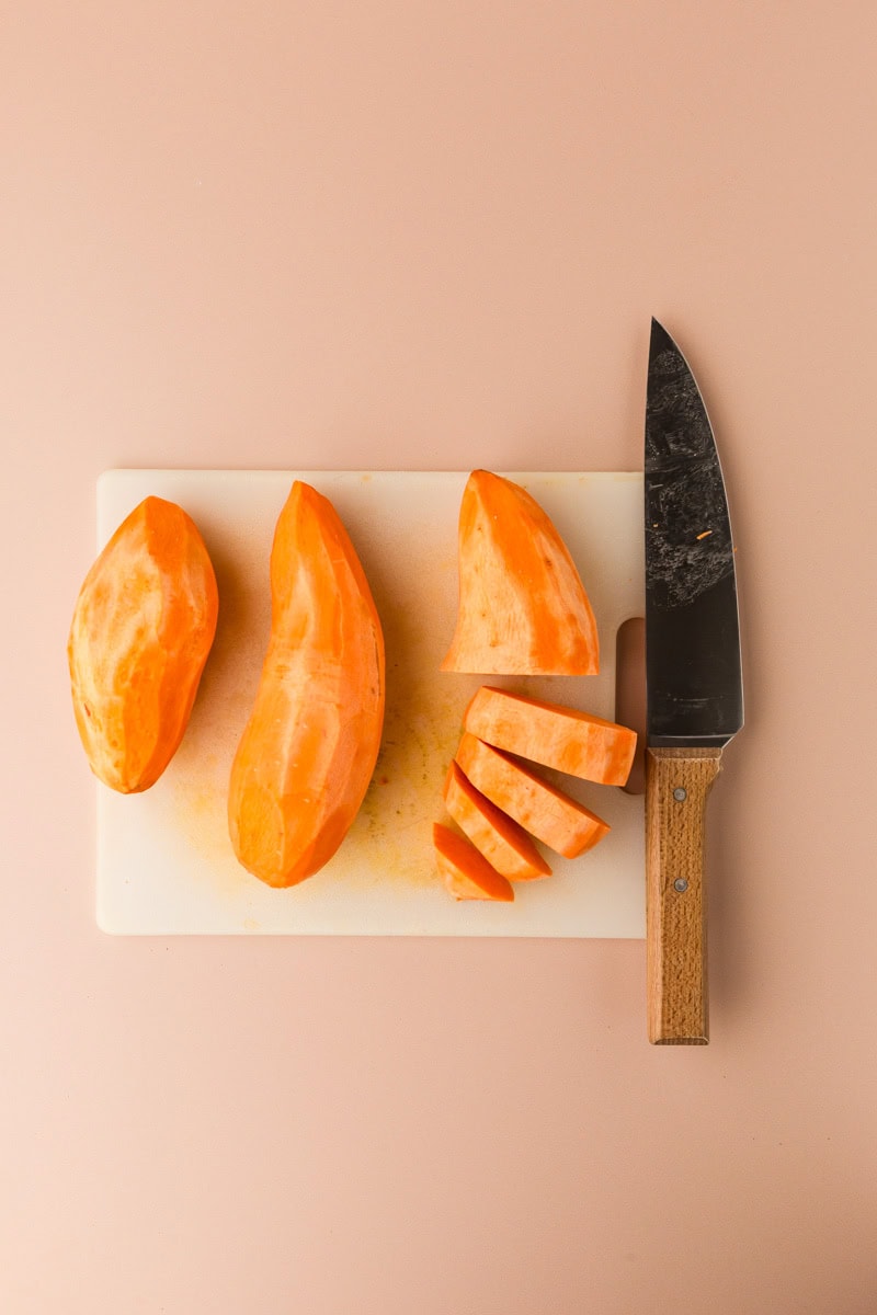 A cutting board with a large kitchen knife and a peeled sweet potato, partially sliced into thick pieces, rests on a beige background. Its the perfect start for a rosemary-infused sweet potato mash.