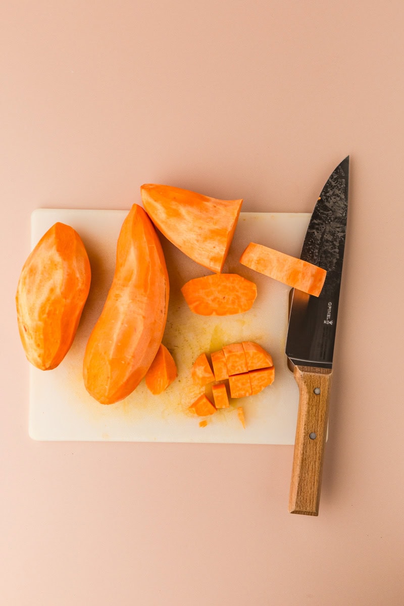 Sweet potatoes, some whole and some diced, are displayed on a white cutting board next to a wooden-handled knife, ready to be transformed into rosemary sweet potato mash. The background is a light beige surface.