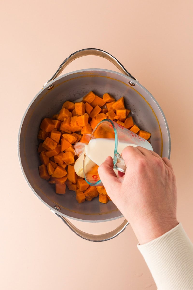 A hand pours cream from a glass measuring cup into a pot filled with diced sweet potatoes, destined to become a creamy rosemary sweet potato mash, against a light pink background.
