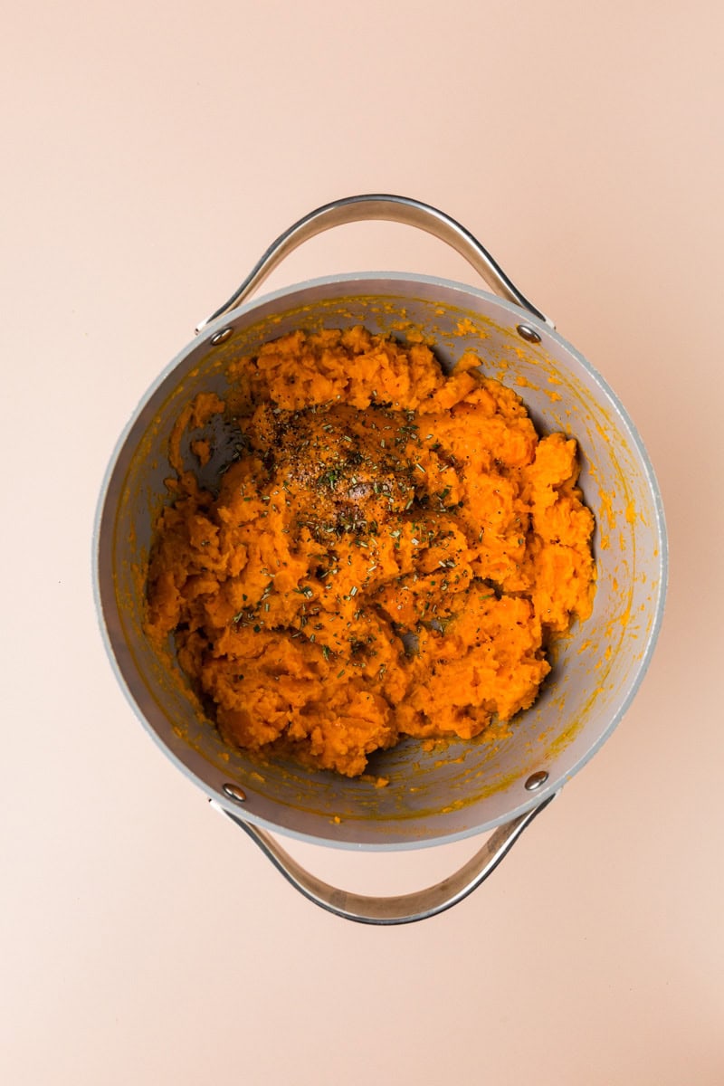 A bowl of rosemary sweet potato mash topped with herbs sits invitingly in a metal pot with two handles, set against a plain background.