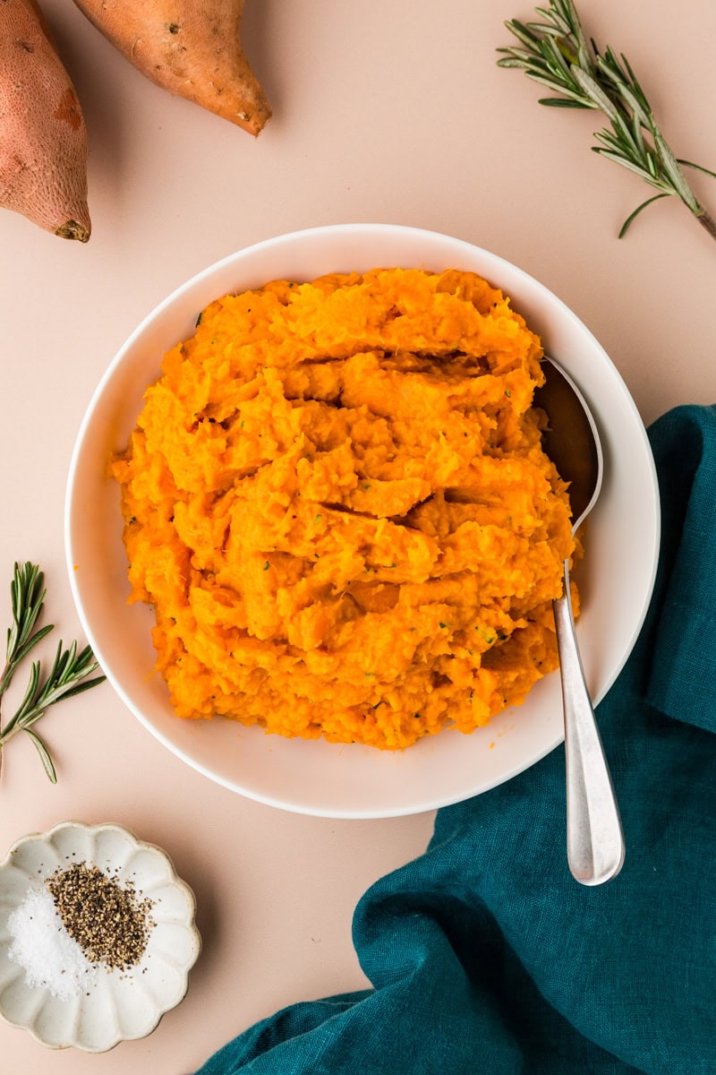 A bowl of rosemary sweet potato mash sits on a neutral table, with a spoon poised at the ready. Sprigs of rosemary, whole sweet potatoes, a teal napkin, and a small dish of salt and pepper elegantly frame the scene.